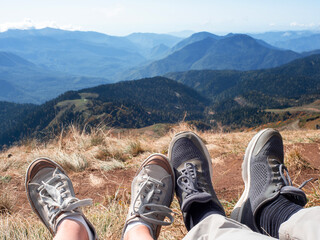 Go to the mountains together. A couple of travelers sat down to relax on the mountain, a first-person view of the mountains, feet in sneakers in the foreground.