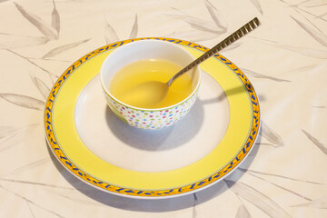 A spoon in delicious fat-free vegetable broth on a round bowl and plate on a floral patterned tablecloth.