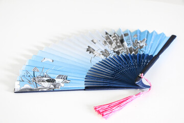 Blue Chinese oriental style fan (fengshan) with pink tassels lying white table background. Images of nature, butterflies over flowers on the fan. Useful for hot weather.