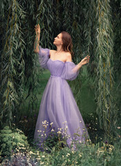 A girl in a lilac long dress stands among the branches of a willow, near a tree. She looks at the...