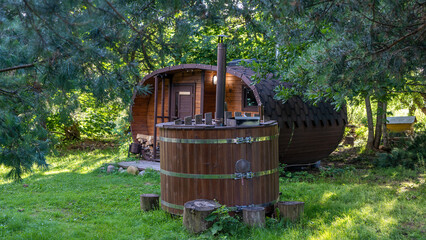 Outdoor wooden barrel sauna in the garden. In foreground wooden bathtub with fireplace. Wooden hot...