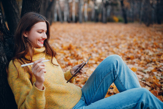 Young woman model sitting with tablet pc and painting with pencil stylus in autumn park with yellow foliage maple leaves.