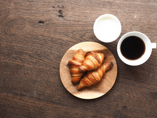 Top view small french butter croissants on round wooden plate served with cup of hot black coffee and glass of milk on wooden background