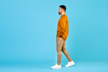 Full length side profile of young male walking, stepping, looking at empty space isolated on blue color background