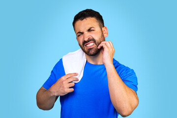  A bearded caucasian man in a blue t-shirt with a towel scratches his beard on a blue background....