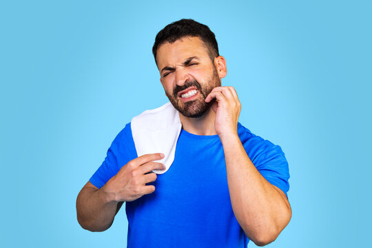  A bearded caucasian man in a blue t-shirt with a towel scratches his beard on a blue background. Irritating itching.