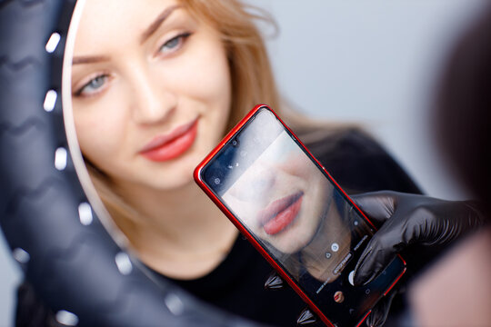 Cosmetologist making photo of work on mobile phone red permanent make up tattoo on young woman lips.