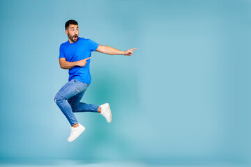 Fototapeta na wymiar Full length portrait of cheerful young man jumping in blue t-shirt pointing finger to the side celebrating success against blue background