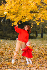 A happy smiling young mother and a little daughter, a child in warm bright clothes, walk together, throw leaves and laugh in an autumn park in nature in the fall in the outdoor forest