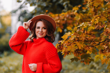 Portrait of a smiling young woman with red hair in a fashionable hat and a red knitted sweater holding autumn leaves while walking in the park outdoors in fall. Selective focus