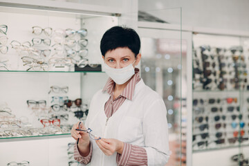Happy Optician in Medical Face Mask Choosing Eyeglasses, female optometrist with glasses.