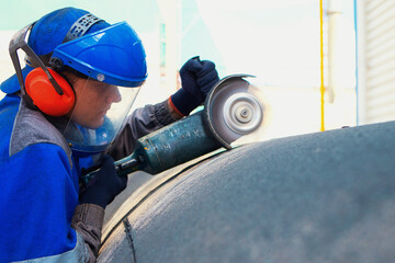 Linear fitter or pipeliner removes plastic insulation from large-diameter pipe with angle grinder. Young worker in overalls and protective mask works outside and processes metal pipe.