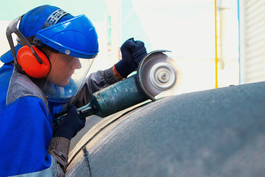 Linear fitter or pipeliner removes plastic insulation from large-diameter pipe with angle grinder. Young worker in overalls and protective mask works outside and processes metal pipe.