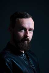 Obraz premium dramatic portrait of bearded millennial guy in leather jacket on dark background