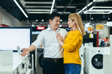 Happy diverse young family man and woman choose electronic household appliances, shop for new apartment in supermarket