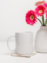 Mockup mug with pink gerbera flowers in vase at the background. Mug for logo, design and text