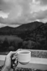 Hand holding a hot latte coffee in a white cup over a blurry background of a landscape with forest, mountains, cloudy sky and a terrace with stones (in black and white)
