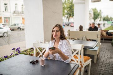 A young attractive girl is holding a smartphone in her hands, checking social communication in a cafe on the street. A woman in casual clothes enjoys using technology during a refreshing cocktail