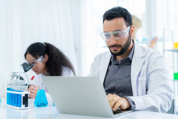 Scientist in lab coat and safety glasses holding flask with blue solution at modern laboratory. Take note on the laptop for analysis. Research development, bio chemistry, medical, pharmaceutical