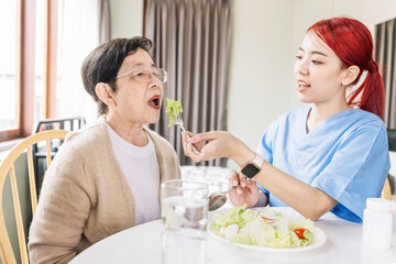 Fototapeta na wymiar Asian woman caregiver wearing medical scrub takes care of senior Asian woman by feeding vegetarian salad to eat at home. caregivers visit home Home health care concept and nursing home.