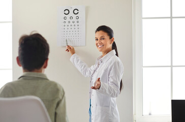Friendly doctor doing eye exam. Professional ophthalmologist testing child's eyesight. Happy young woman in white coat standing in medical office, pointing at eye chart and smiling
