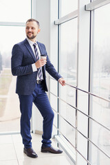 A young man in a business suit with a cup of coffee stands at a large window and looks out. Young businessman on the break