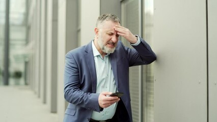 Mature business man with dizziness, vertigo a severe headache standing on a city street leaning against a wall. outside. Senior male with high blood pressure or stroke holding head massaging. Outdoor