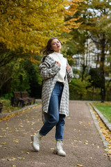 Fototapeta premium European elegant young woman in a stylish trench coat in a white sweater and blue jeans walking in a park. warm autumn day.