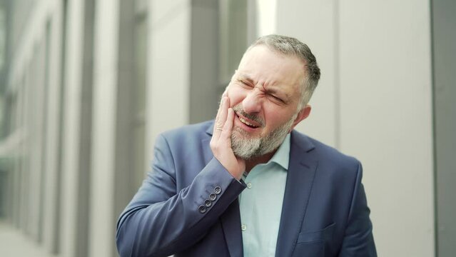 Mature Business Man With Severe Toothache Standing On A City Street Near An Office Building. Gray Haired The Employee Having Dental Pain Referred.diseases Standing Front Office Building Outside