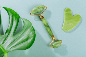Green jade face roller and gua sha scraper with leaves on light blue background. Massage tool for...