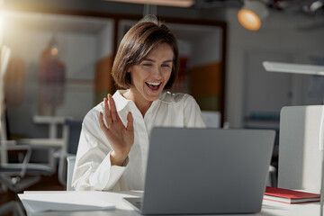 Businesswoman making videocall to friends and waving hi sitting in modern office. High quality photo