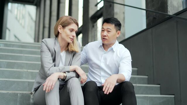 Asian male employee supports upset depressed female co-worker colleague. Sitting on the steps stairs outside an office building. young man comforting sad woman at work outdoors 