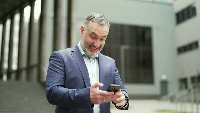 Mature business man employee entrepreneur looking phone smartphone with shouts win gesture, read good news, investment, profit growth. Happy male investor rejoices celebrate victory won outside office
