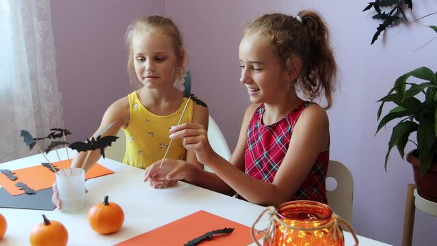a selective focus of two funny and cute schoolgirls, they are sitting at a table, looking at each other and smiling, making decorations for the Halloween holiday
