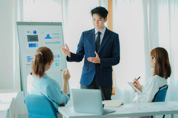 Successful Asian man, manager, businessman working with an Asian female colleague at a desk in a company office with a financial graph and laptop on the table.