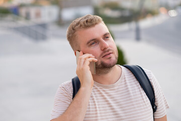 Handsome blond bearded smiling man with casual clothes,having call using smartphone in city outdoors.Happy caucasian guy wearing backpack,speaking,using phone.