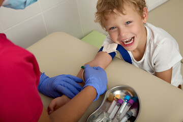 Cute cheerful boy boldly extended his hand to a nurse in a hospital laboratory to donate blood from a vein for analysis. A nurse in a children's polyclinic takes blood from a child from a vein