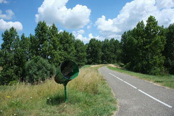 Bicycle path with a metal garbage bin for cyclists between uncultivated farmland and trees in the background, summer day with a sky filled with clouds, in the Netherlands Holland - obrazy, fototapety, plakaty