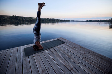 A yogi woman does a headstand on the dock near the river in nature.