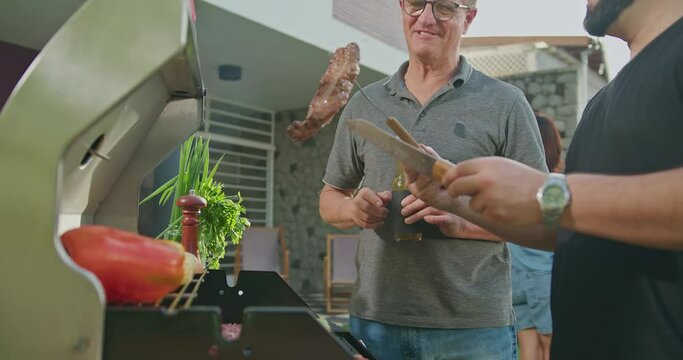 Father standing by son grilling meat. Adult son showing steak to dad. Friends and family gathered for barbecue. Parent talking to son during summer holiday celebration