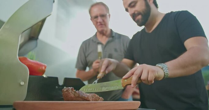 Man cutting meat at barbecue. Chef preparing food for family and friends. Father holding bottle of beer observing adult son prepare meal at home backyard grill