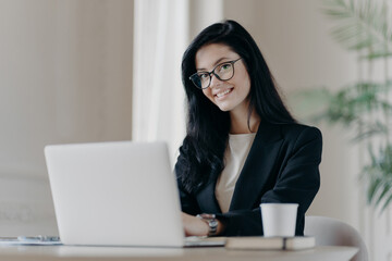 Positive dark haired woman makes research, browses information on laptop computer, connected to wireless internet, being happy wears formal clothes and spectacles drinks coffee. Economist works online