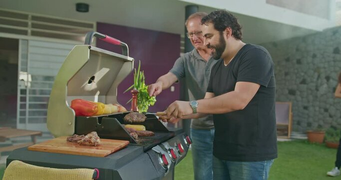Barbecue chef preparing food at grill in garden backyard party. Father observing adult son prepare meat