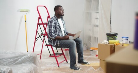 Man of African American sits on ladder in apartment on knees holding laptop looking for inspiration...