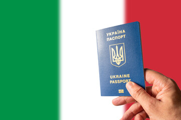 Ukrainian biometric passport in the hand of a person against the background of the national flag of Italy. Emigration, business, tourism, flight from the war in Ukraine to Italy.