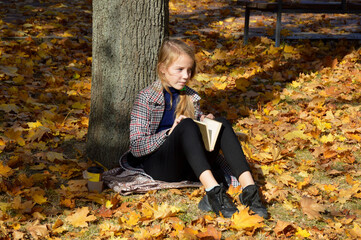 a beautiful girl in a plaid coat stands leaning on a tree trunk in an autumn park. holds a bouquet of yellow maple leaves in her hands and looks intently to the side