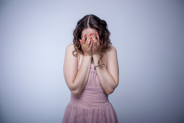 Fototapeta premium A young girl in a beautiful dress stands on a white background and covers her face with her hands. The girl is upset or crying.
