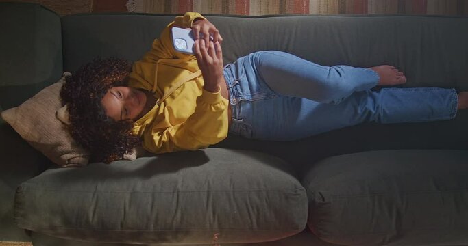 African American woman lying on sofa holding smartphone seen from above angle. A black girl taking selfie with phone