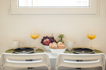 Small white folding table under a window with breakfast service, glasses with orange juice, a bowl...