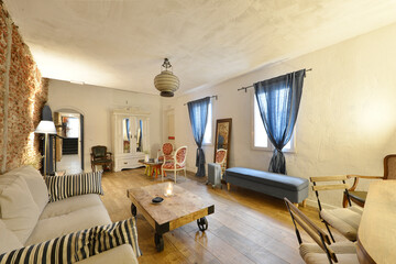 Living room with assorted chairs and armchairs, wooden floors, windows with blue curtains, exposed brick wall and wooden furniture